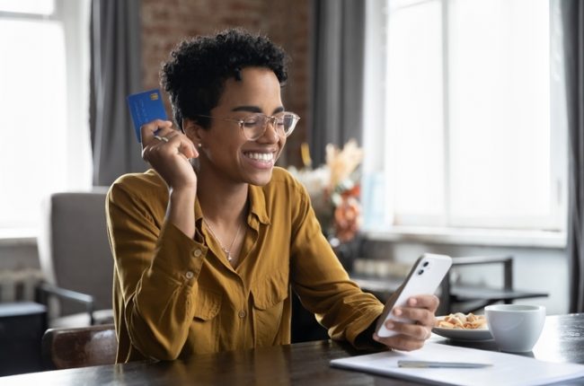 African,Woman,Sit,At,Desk,Hold,Credit,Card,Use,Smartphone