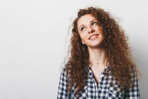 iStock 934915908 295x197 - Dreamy curly woman with attractive look, dressed in checkered shirt, poses against white background, thinks about something pleasant. Good looking young female model being deep in thoughts